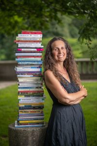 Lisa Tener leans against a multicolored stack of books in front of a green park background